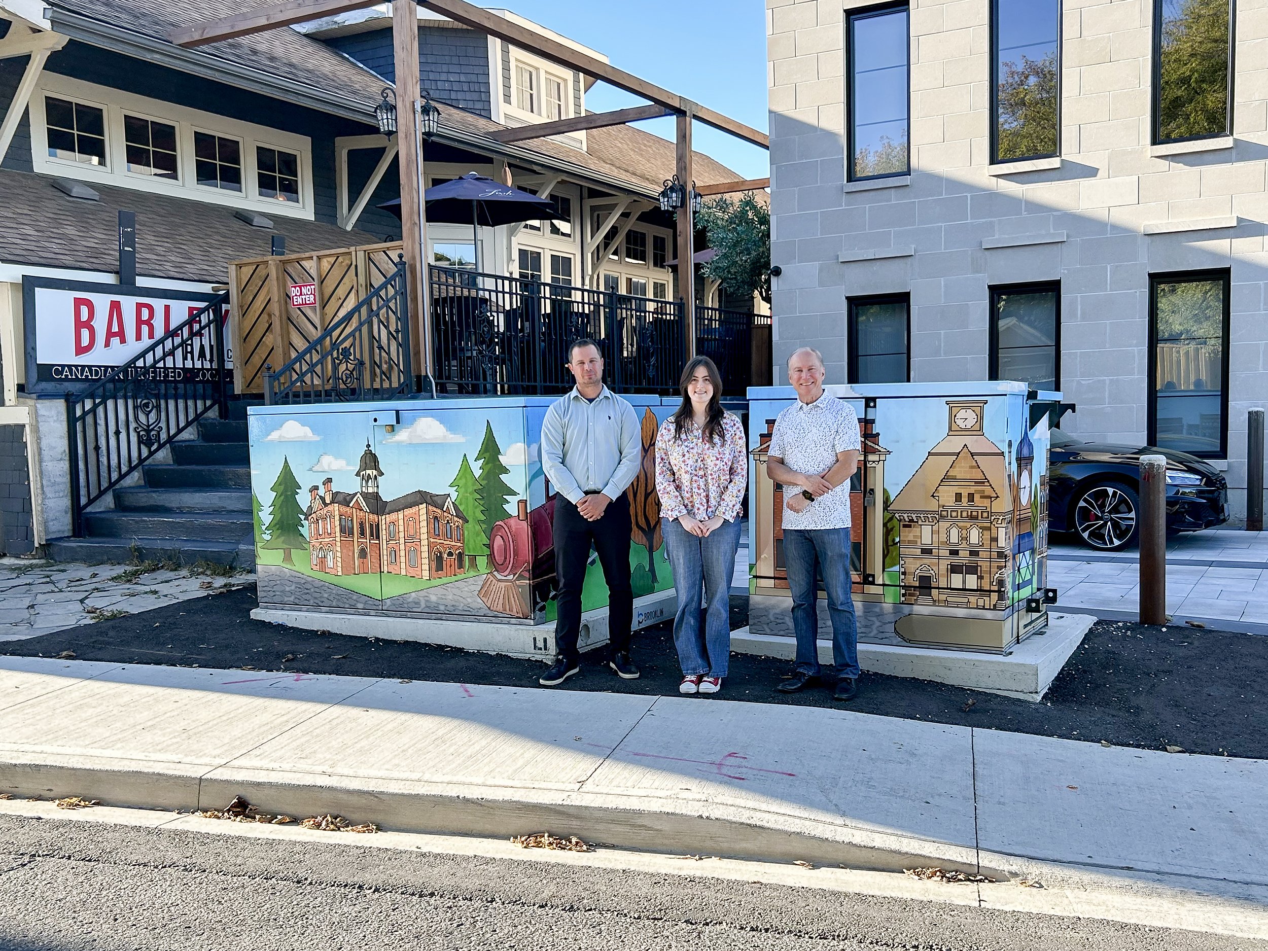 Three people stand in front of two utility boxes with a vinyl wrap of pixel art on the boxes.